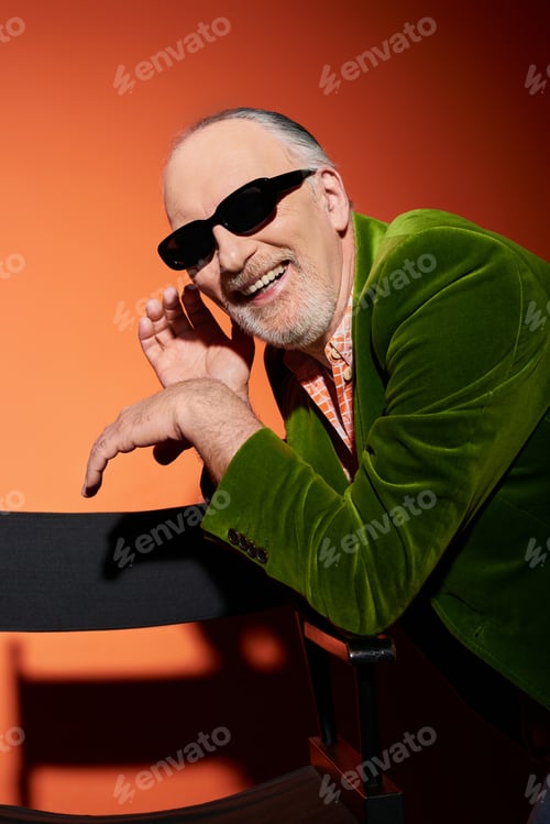 Preview: carefree and excited bearded man in stylish outfit smiling near chair on red and orange background