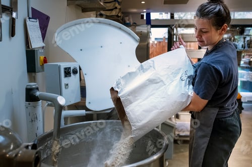 Preview: Woman wearing apron standing in an artisan bakery, pouring flour into industrial mixer.