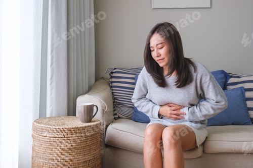 Preview: A sick woman suffering from stomachache, abdominal pain while sitting on sofa at home