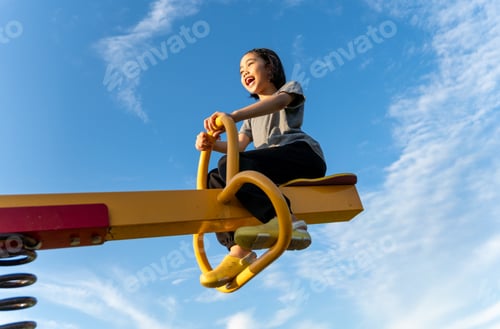 Preview: A happy young Asian girl on a yellow seesaw at a playground.