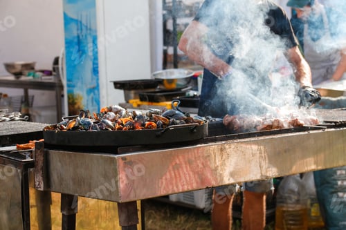 Preview: man cooking bbq meat at festival outdoor. Seafood paella. Chef grilling sausages in park outside. Co