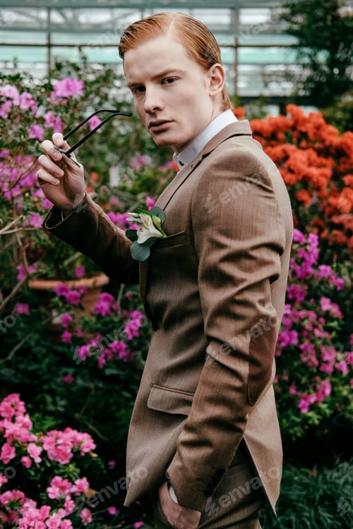 Preview: side view of stylish young man with red hair and eyeglasses in hand standing in glasshouse