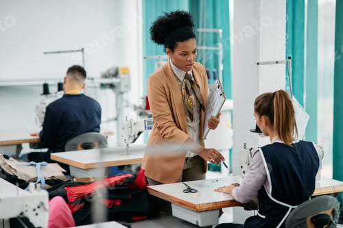 Preview: Black clothing factory designer talking to a seamstress at production line.