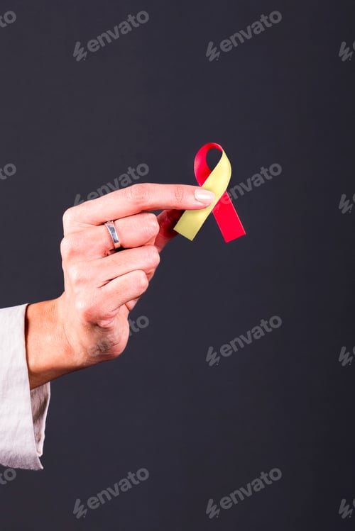 Preview: Woman’s hand holding a red and yellow ribbon, symbol of COVID-19 and cultural diversity awareness