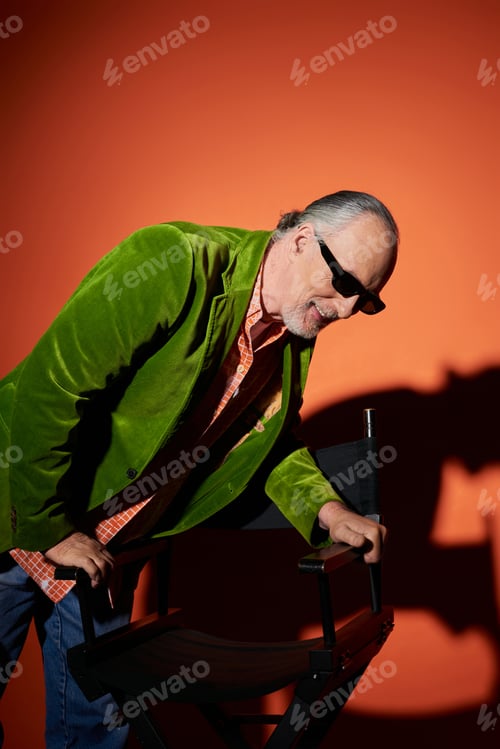 Preview: pleased senior man in stylish outfit laughing while standing near chair, dark sunglasses, green