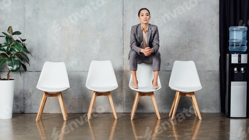 Preview: serious asian businesswoman in formal wear sitting on chair and looking at camera in waiting hall