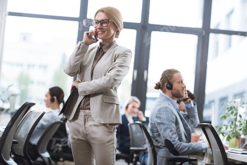 Preview: selective focus of smiling businesswoman in suit with notepad talking on smartphone while standing