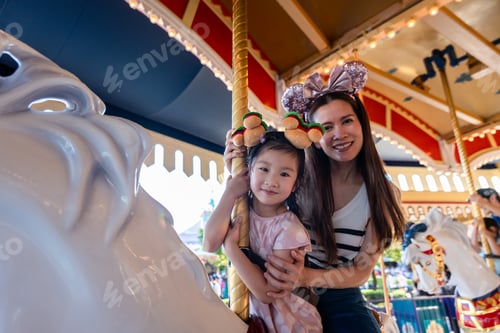 Preview: Asian mother and adorable kid daughter riding carousel at theme park.