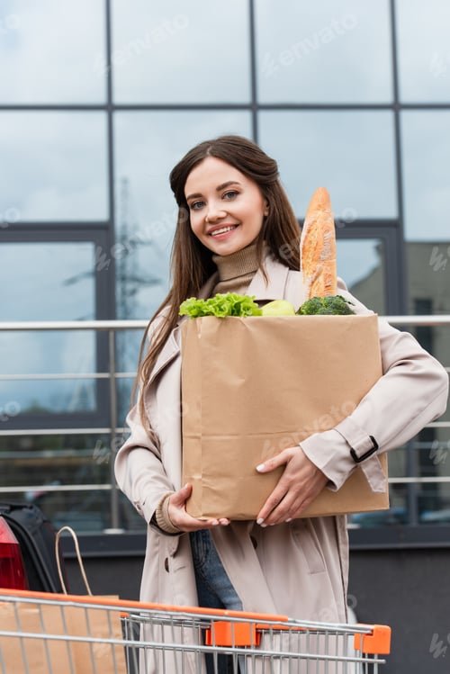 Preview: happy woman looking at camera while holding shopping bag with fresh food outdoors