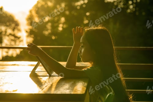 Preview: Little girl using tablet pad while sitting in outdoor during sunrise.