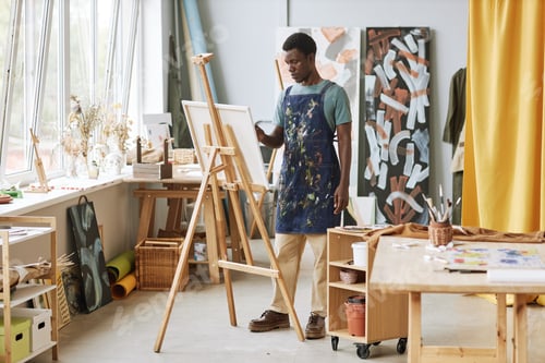 Preview: Young serious creative male artist in apron standing in front of easel