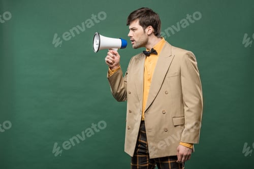 Preview: handsome man in vintage clothes talking in loudspeaker isolated on green with copy space