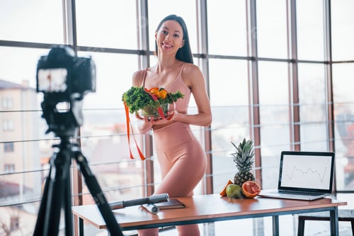 Preview: Young fitness vlogger doing video indoors by standing near table with healthy food and laptop