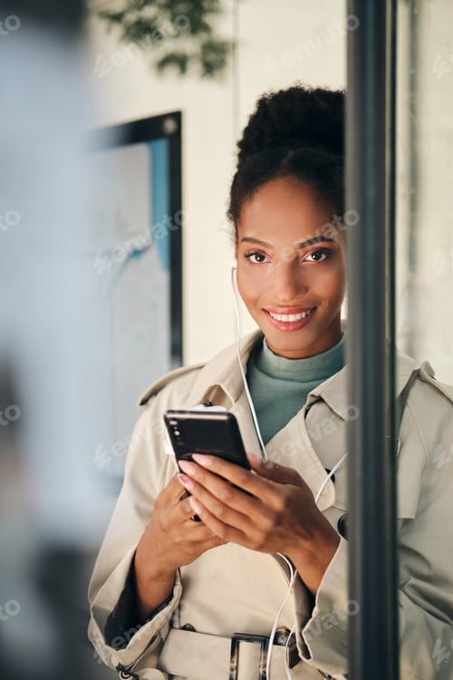 Preview: Beautiful African American girl in stylish trench coat with cellphone looking in camera at bus stop