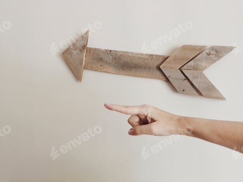 Preview: Hand pointing to left in front of wooden arrow decor on plain white wall