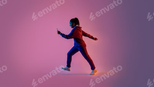 Preview: Black lady in sportswear walking and using cellphone over pink neon background, full body, side view