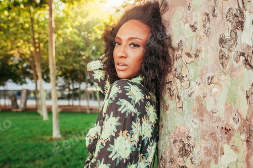Preview: Thoughtful young afro woman leans on tree outdoors