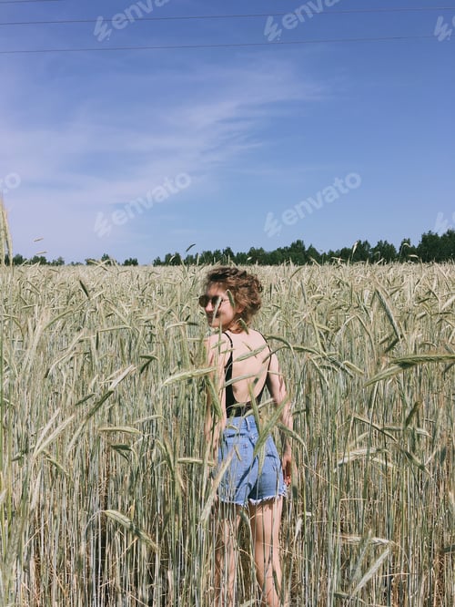 Preview: A young girl in shorts and a top with an open back stands in a wheat field. Summertime. Hot days
