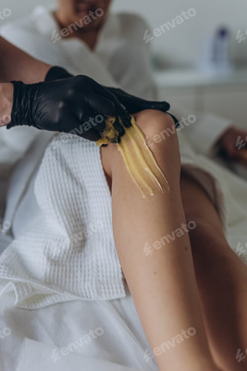 Preview: Beautician depilating young woman's legs with liquid sugar in spa center