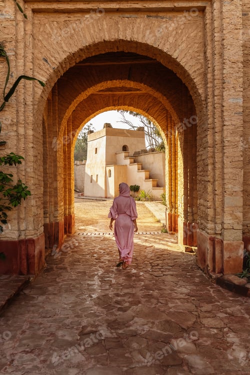Preview: Woman in Pink Traditional Clothing Walking Through Historic Stone Archway in Marrakesh Medina