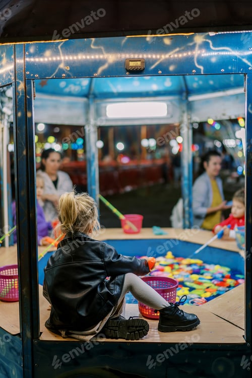 Preview: Little girl catches fish with a toy fishing rod while sitting by a toy pool at a fair with children