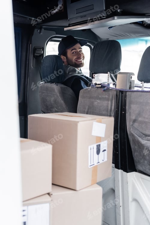 Preview: Smiling delivery man looking at camera near packages in car