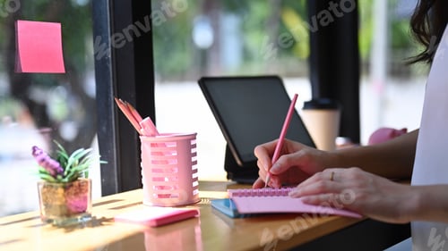 Preview: Cropped shot of female designer writing on notebook while sitting near window in office.