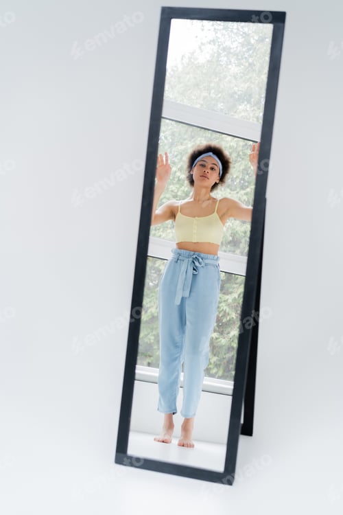 Preview: Barefoot african american woman in headband looking at mirror on grey background