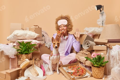 Preview: Indoor shot of happy woman eats pizza waves hello with palm sits crosed legs near dog surrounded by