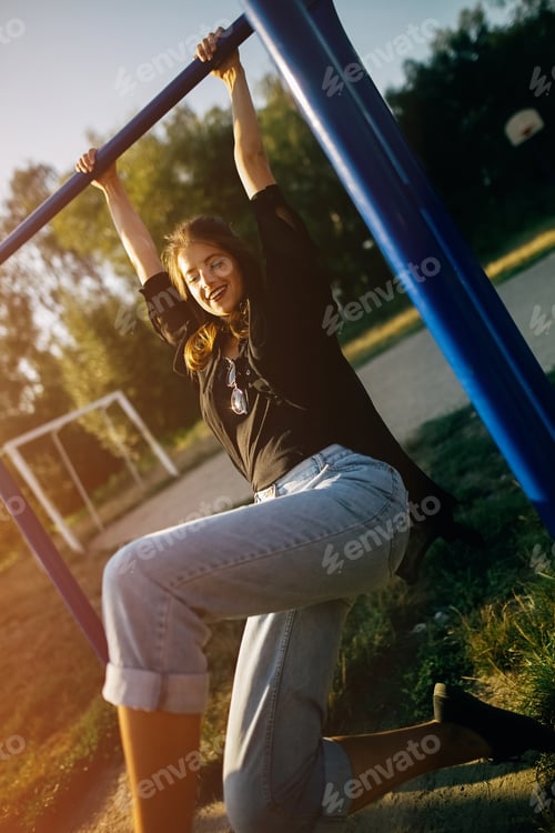 Preview: Fashionable cool woman playing and hanging on horizontal bars in evening