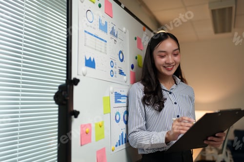 Preview: Young professional woman reviewing data on charts displayed on a whiteboard, smiling and taking note