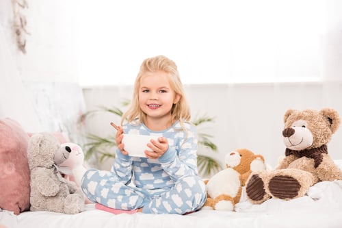 Preview: smiling child sitting in bed with soft toys and holding bowl with breakfast