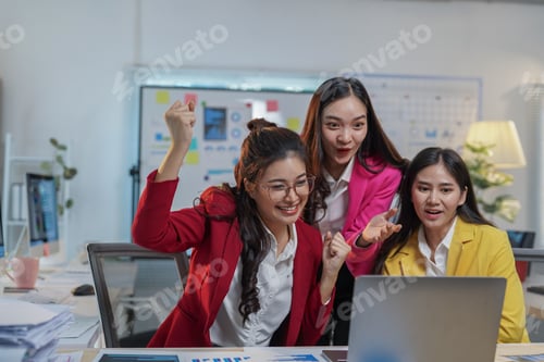 Preview: Three young asian businesswomen celebrating success while looking at a laptop screen, raising their