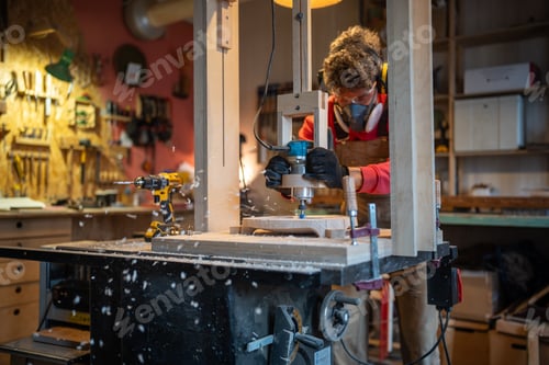Preview: Man carpenter in noise-cancelling headphone, glasses, respirator milling wooden plank in joinery