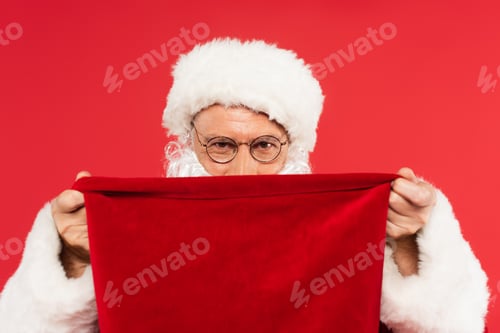 Preview: Santa claus in eyeglasses covering face with sack isolated on red