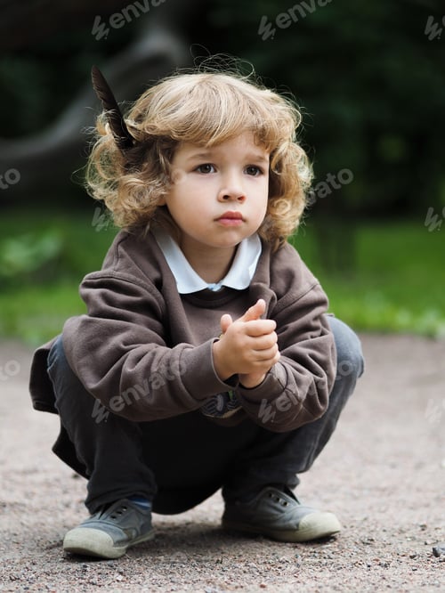Preview: Portrait of a cute curly-haired boy with a feather behind his ear sitting on the street.