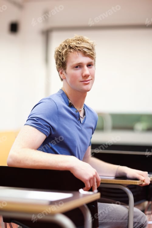 Preview: Man in his twenties sitting at classroom desk in lecture room wearing blue henley, earbud visible