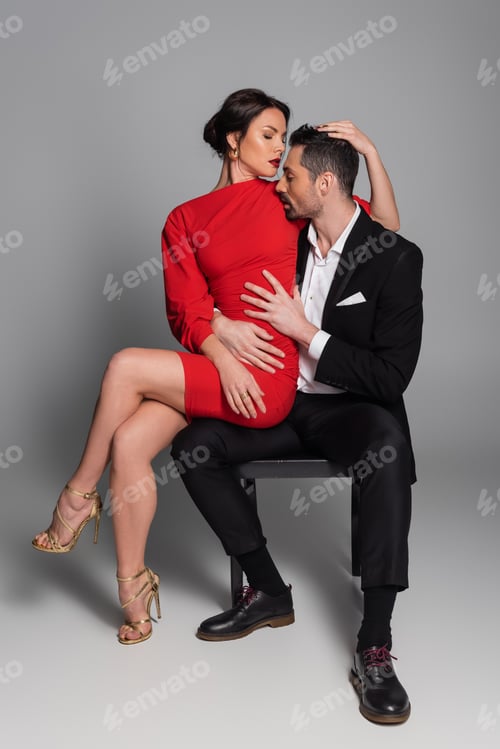 Preview: Full length of seductive woman in red dress touching boyfriend while sitting on chair on grey