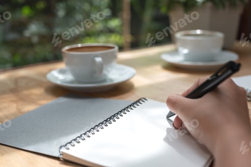 Preview: Closeup image of a hand writing down on a white blank notebook with coffee cup on wooden table