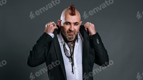 Preview: elegantly dressed punk man lifts lapels of his jacket looking defiantly at camera in studio shot