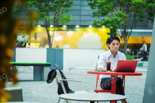 Preview: Businessman wearing white shirt sitting outdoors at red table, working on laptop.