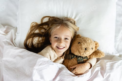 Preview: Cute little Caucasian girl with blonde hair wearing pajamas in bed with teddy bear
