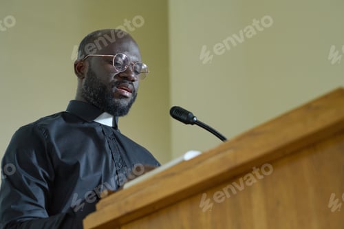Preview: Young priest in black shirt with clerical collar pronouncing speech by pulpit
