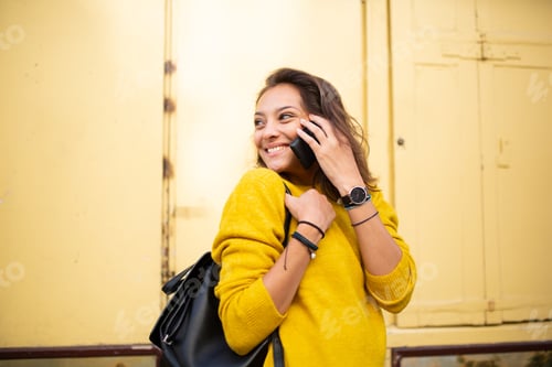 Preview: Side of smiling young woman talking with phone outside and looking over shoulder