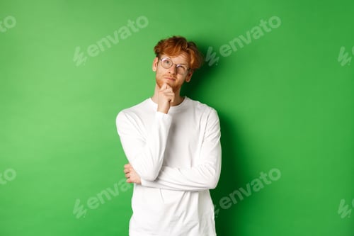 Preview: Thoughtful redhead man in glasses making choice, looking up and thinking, standing over green