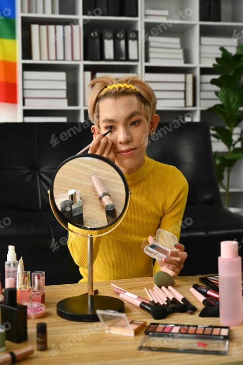 Preview: Young gay man doing daily makeup in living room with rainbow flag on background.