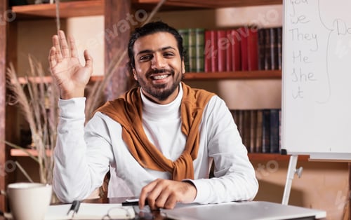 Preview: Joyful Indian Teacher Man Waving Hello Posing Sitting In Classroom