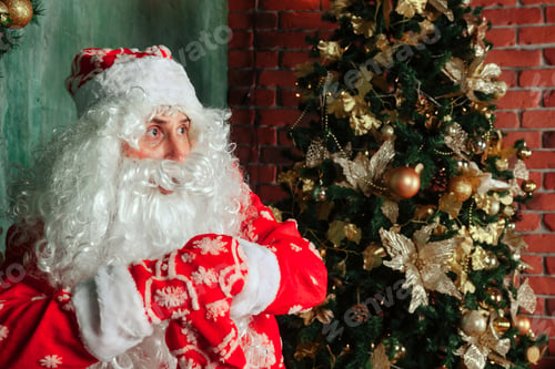 Preview: Santa Claus with bag of gifts by Christmas fireplace in decorated living room with Christmas tree