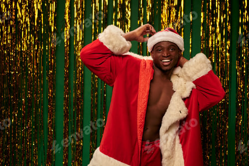 Preview: cheerful african american guy posing in santa costume on shirtless body near shiny tinsel on green