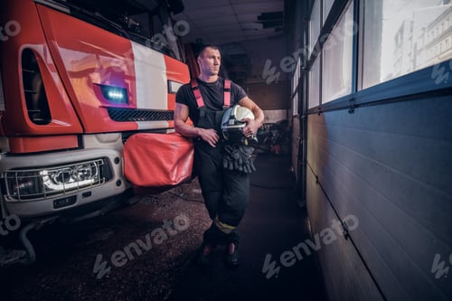 Preview: Fireman wearing protective uniform standing next to a fire engine in a garage of a fire department